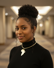 A woman wearing a black turtleneck with a necklace featuring an African map design in a blurred indoor setting.