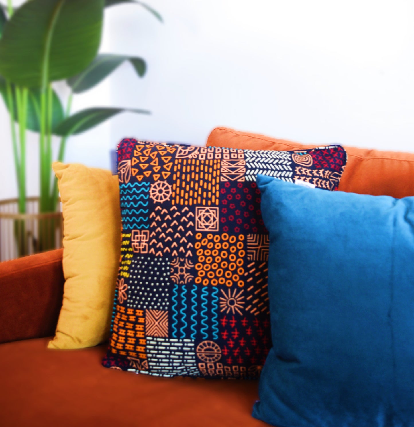 A colorfully patterned and designed pillow cover on an orange couch. The design contains colors such as blue, magenta, peach, orange, and yellow.