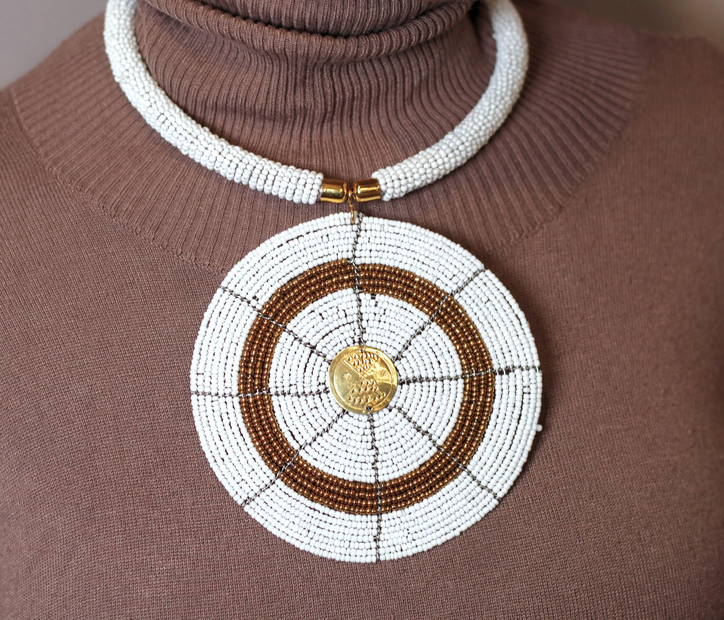 A person wearing a brown top and the Karani Beaded Necklace which is predominantly white with a circular pendant featuring a mix of gold and white beads.