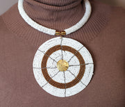 A person wearing a brown top and the Karani Beaded Necklace which is predominantly white with a circular pendant featuring a mix of gold and white beads.