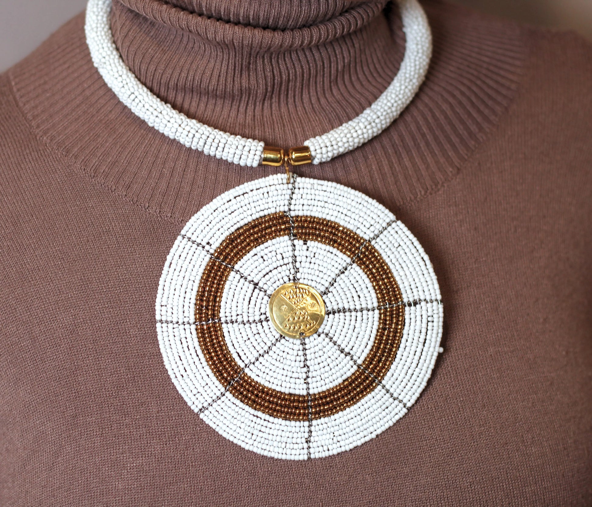 A person wearing a brown top and the Karani Beaded Necklace which is predominantly white with a circular pendant featuring a mix of gold and white beads.