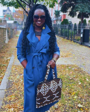 Woman in a blue coat holding a patterned bag on a sidewalk with autumn leaves.