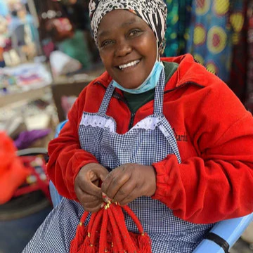 Woman in a red jacket and checkered apron holding red beaded neckalces in a market setting
